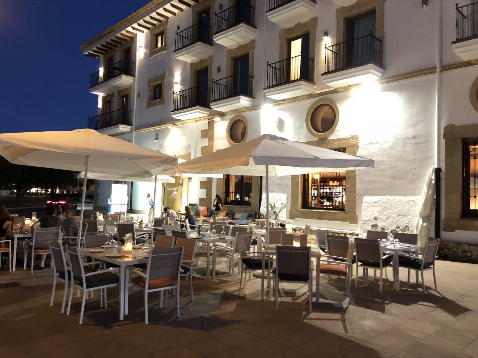 Un área de comedor al aire libre con mesas y sillas, sombrillas y un edificio con balcones al fondo.