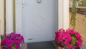 A white door with a diamond-shaped window and a keyhole, flanked by two columns and potted pink flowers.