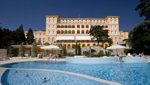 A large pool area with multiple swimming pools and sun loungers, surrounded by a building with multiple balconies and a stone bridge.