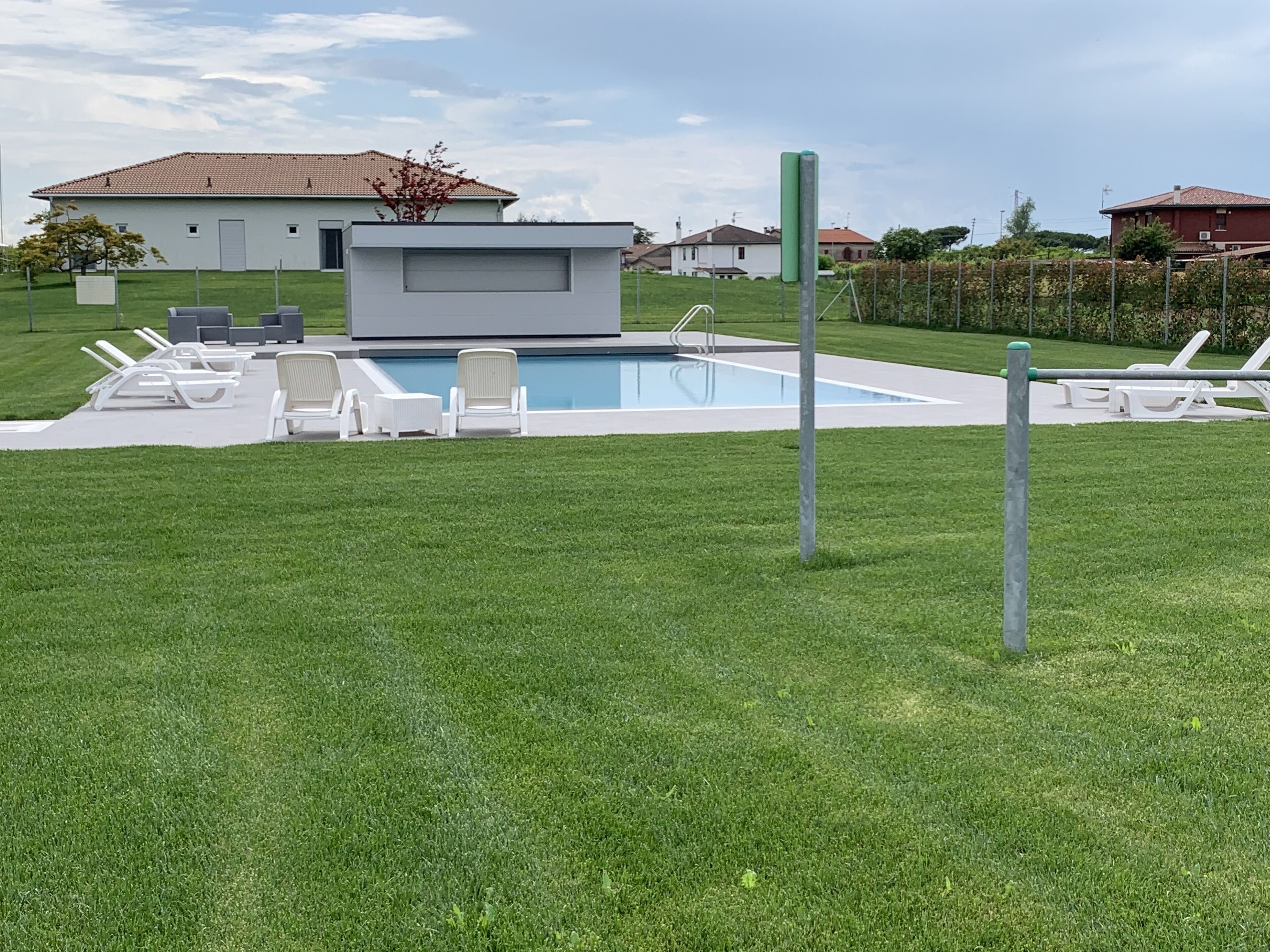 Un área de piscina moderna al aire libre con sillones de descanso y un pequeño edificio, rodeada de césped cuidado y una cerca.