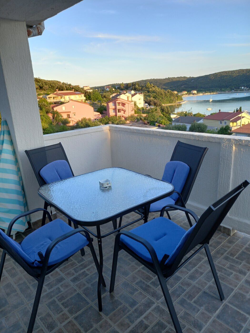 Un balcón con mesa de vidrio y sillas azules, con vistas a una zona residencial y una masa de agua.