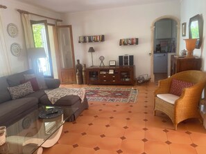 Ein Wohnzimmer mit einer Couch, einem Korbstuhl, einem Glastisch, einem Teppich mit Muster und einem Bücherregal.