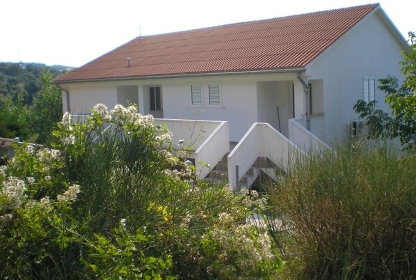 Une maison de plain-pied avec un toit recouvert de tuiles rouges, des murs extérieurs blancs et un petit porche doté d’une balustrade blanche.