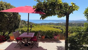 Un patio con mesa y sillas, un paraguas rojo y vistas al mar y las colinas.