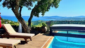 Um deck de madeira com cadeiras de lounge com vista para uma piscina e uma paisagem com mar e colinas.