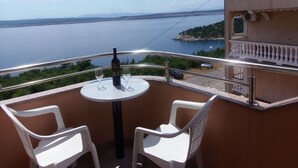 A balcony with two white plastic chairs, a small round table, and a bottle of wine. The balcony overlooks a large body of water and a distant shoreline.