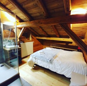 Ein Schlafzimmer im Mansardenbereich mit Holzboden, einem Bett, einer Duschkabine aus Glas sowie einem kleinen Badezimmer mit Waschbecken und Heizkörper.