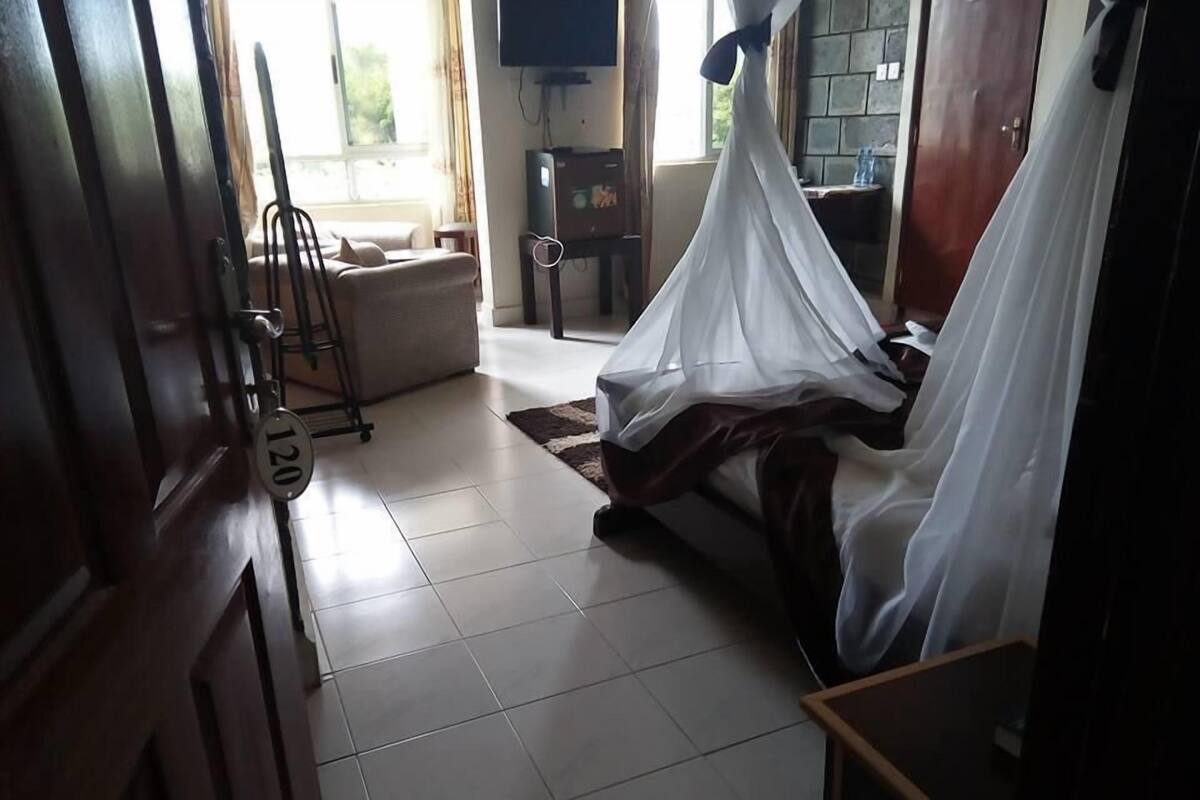 A room with a bed covered in white sheets, a wooden door with a number plate, a small table, and a television on the wall.