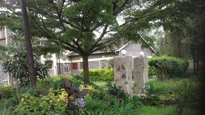 A garden with a stone structure, a tree, and a building in the background. - Lanet Matfam Resort (Nakuru)