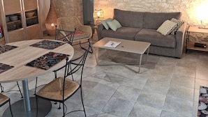 A cozy living room with a stone wall, wooden ceiling beams, a grey sofa, and a round dining table with chairs.