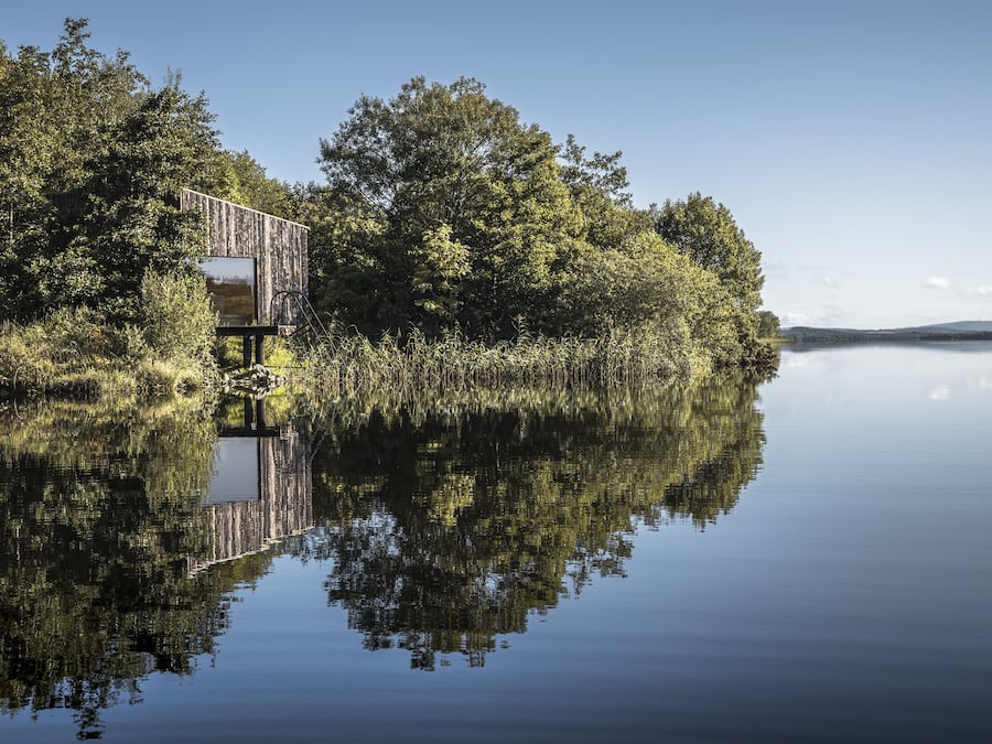 Una cabaña de madera junto a un lago tranquilo rodeada de árboles densos.