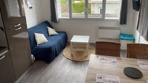 A compact room with a blue sofa, a small white table, wicker chairs, a wooden floor, and a window with curtains. - Quai Ouest: 2/4p rental 100m from Goldbeach beach (Ver-sur-Mer)