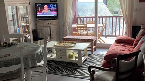 A living room with a television, a dining table, and a sofa. There is a balcony with a view of the outside.