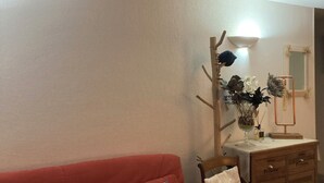 A living room with a red sofa, wooden chair, and a wooden dresser with drawers. There is a wall-mounted lamp and a mirror on the dresser.
