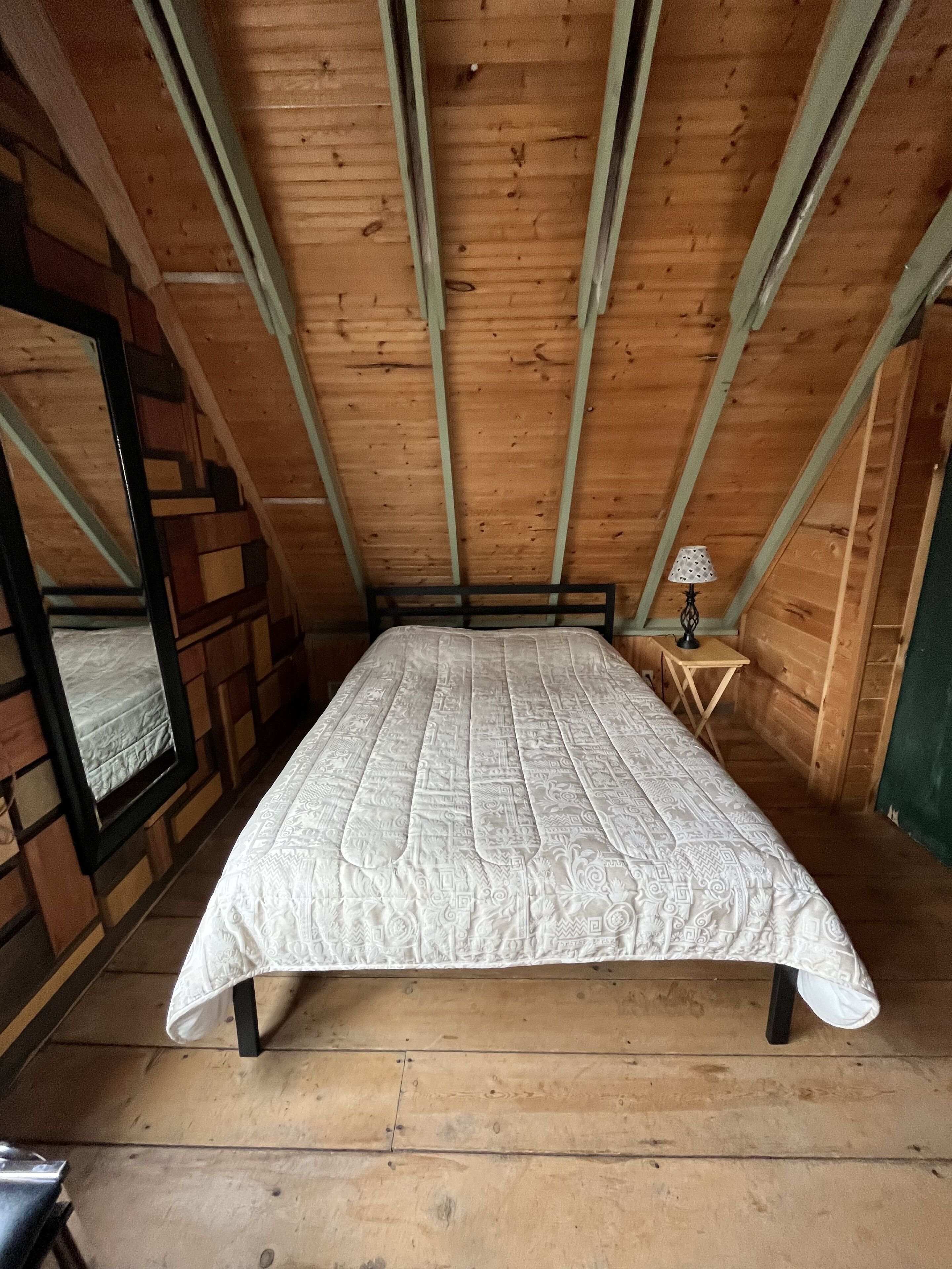 Ein Schlafzimmer mit Holzbalkendecke, einem Bett mit gemustertem Überwurf, einem kleinen Nachttisch mit Lampe und einem Spiegel an der Wand.