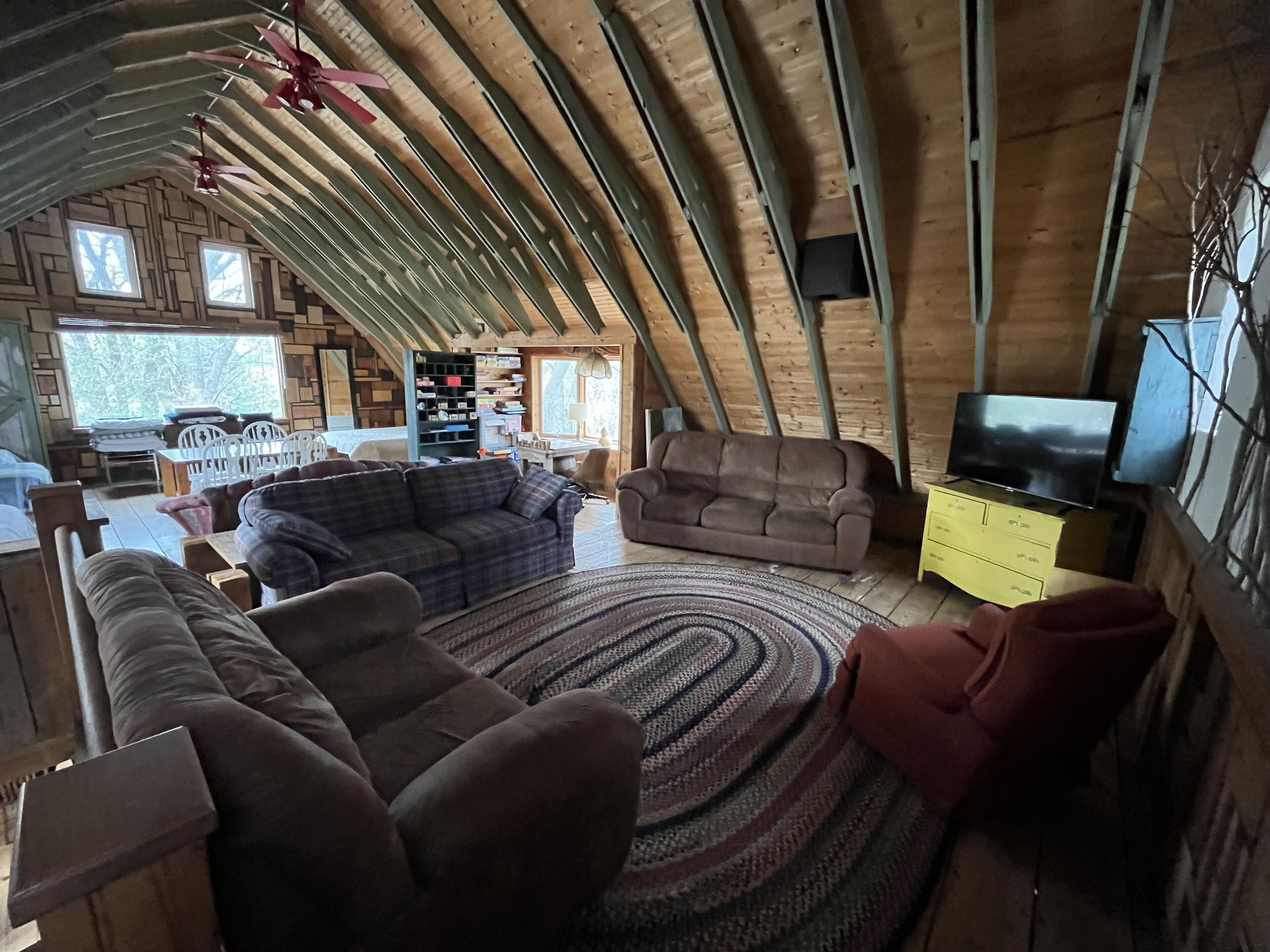 A cozy loft-style living room with a high ceiling, wooden beams, a large window, a sofa, a loveseat, a TV, and a bookshelf.