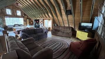 A cozy loft-style living room with a high ceiling, wooden beams, a large window, a sofa, a loveseat, a TV, and a bookshelf.