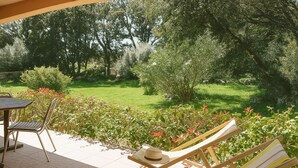 Une terrasse avec une table et des chaises, un fauteuil de relaxation avec un chapeau, et une vue sur un jardin verdoyant avec des arbres.