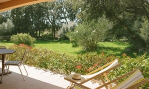 A patio with a table and chairs, a lounge chair with a hat, and a view of a grassy garden with trees.