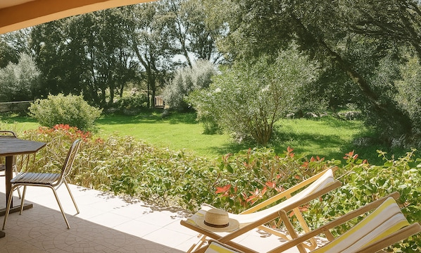 A patio with a table and chairs, a lounge chair with a hat, and a view of a grassy garden with trees.