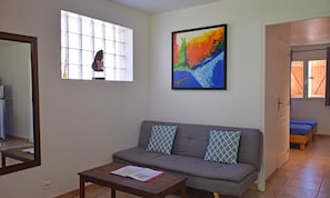 A living room with a grey sofa, two patterned cushions, a wooden coffee table with a magazine, a colorful abstract painting on the wall, a window with a decorative item, and a door leading to another room.