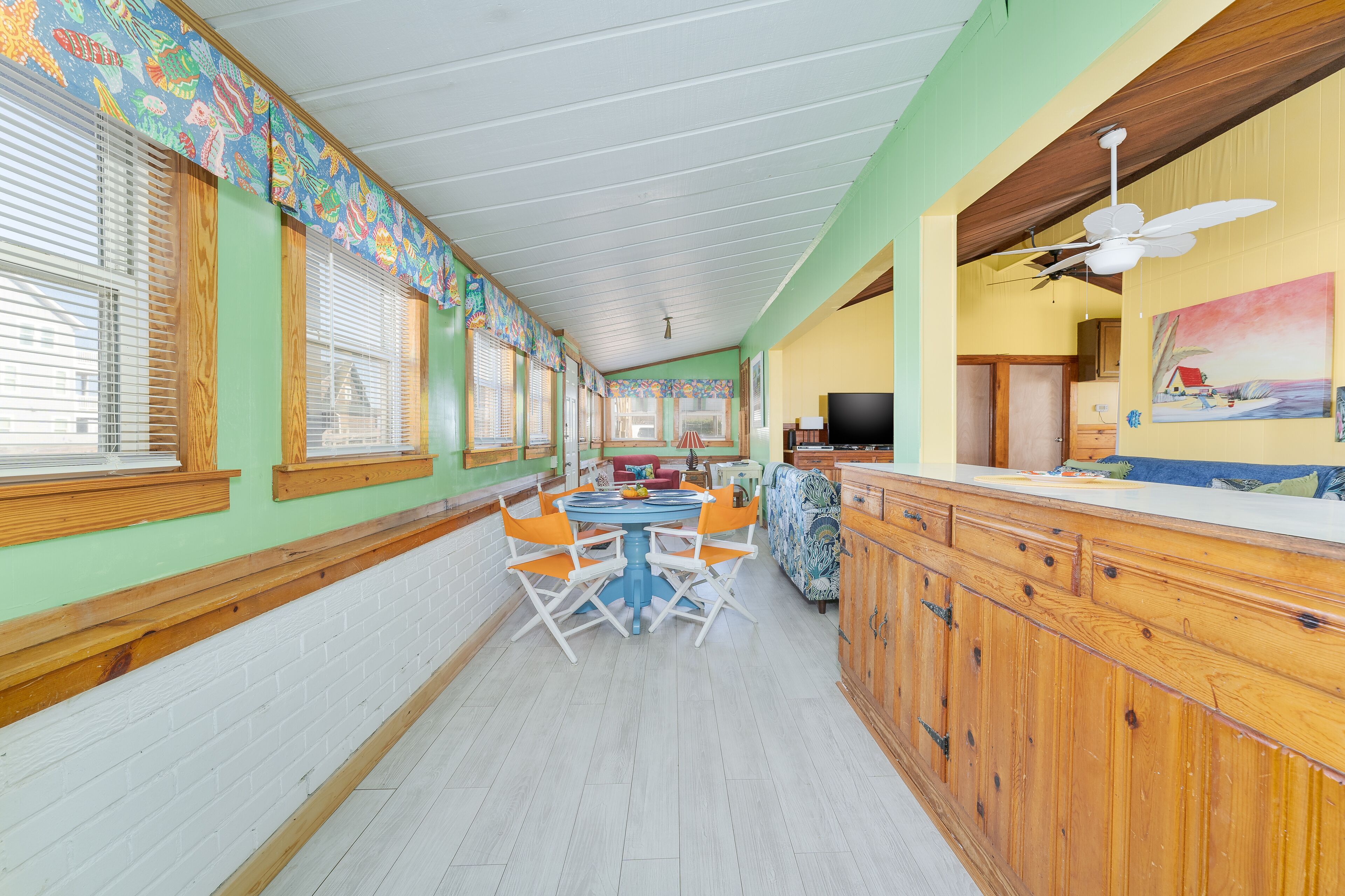 A long dining area with a high ceiling and wooden beams, featuring a central round table with orange chairs and a television on a wooden counter.