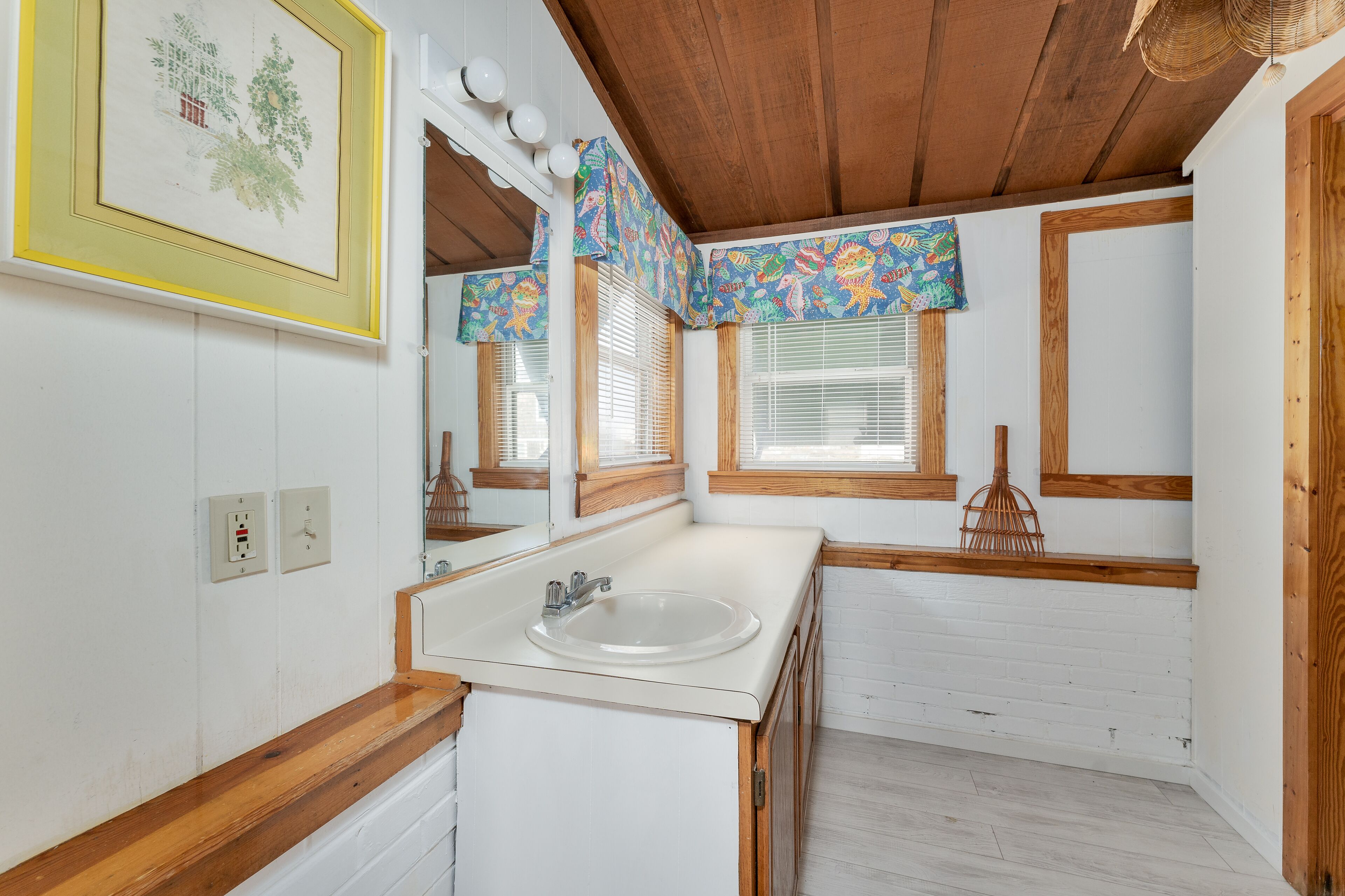 A bathroom with a wooden ceiling, white walls, a sink, and a window with a colorful valance.