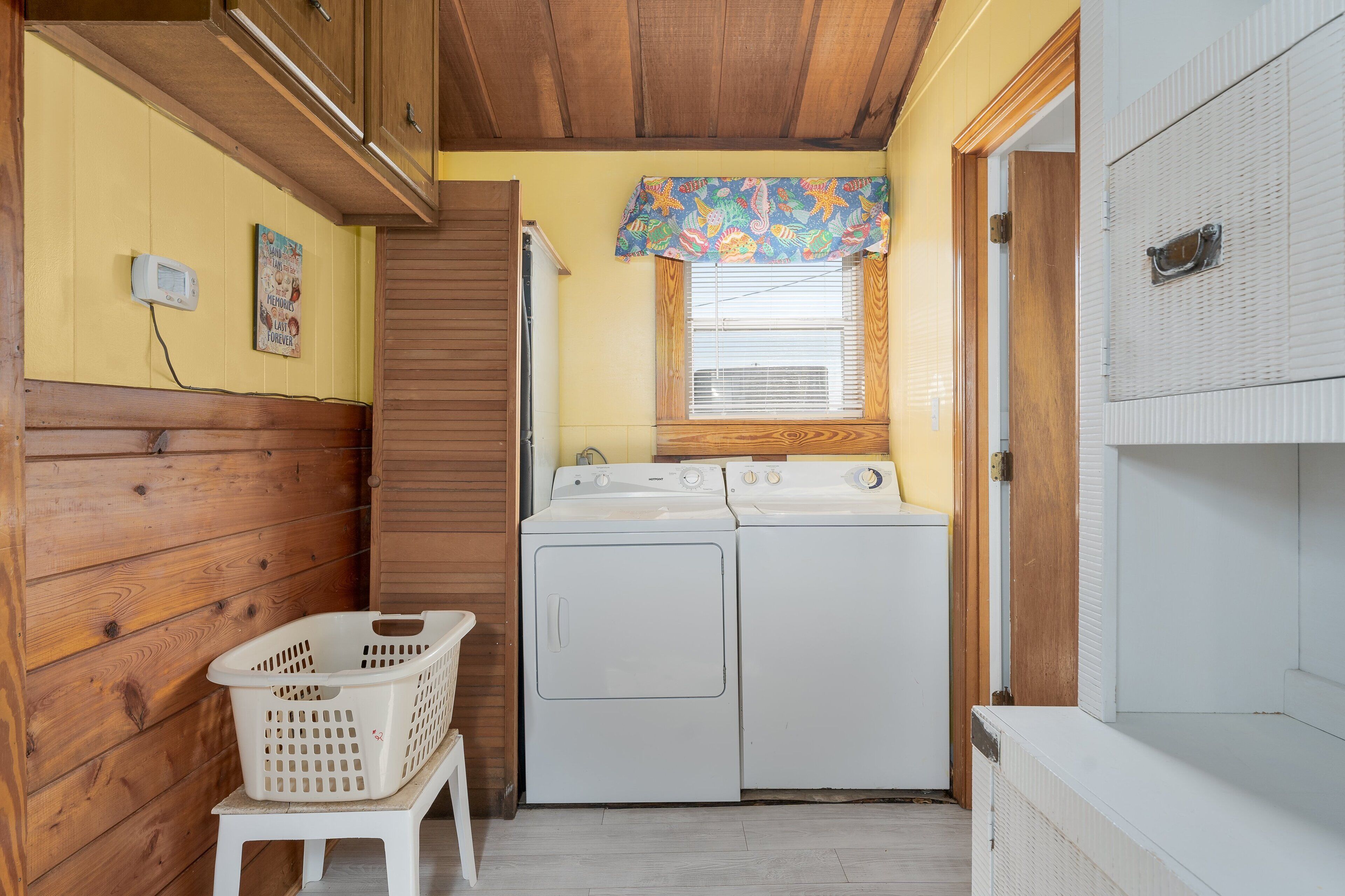 Un lavadero con dos lavadoras blancas, una pared de madera, una pequeña ventana con un adorno colorido y una cesta de ropa blanca sobre un taburete.