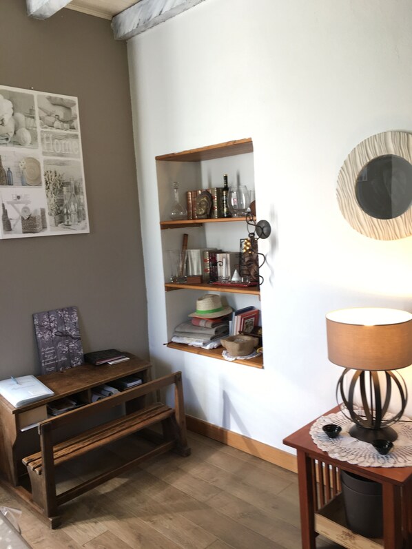 Habitación de esquina con un banco de madera, una mesita con lámpara y un estante en la pared con diversos objetos.