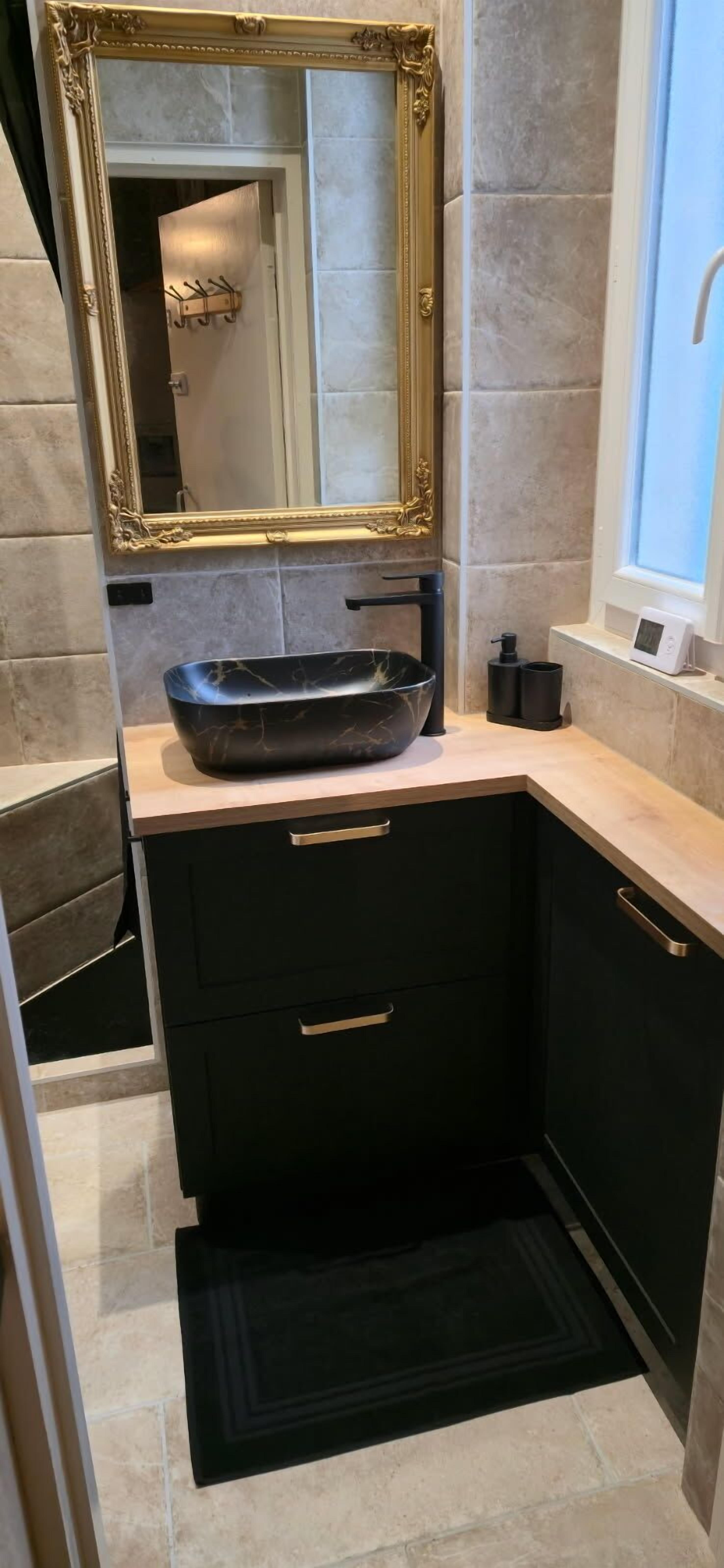 Une salle de bain avec un lavabo noir, un miroir encadré d’or et une fenêtre.