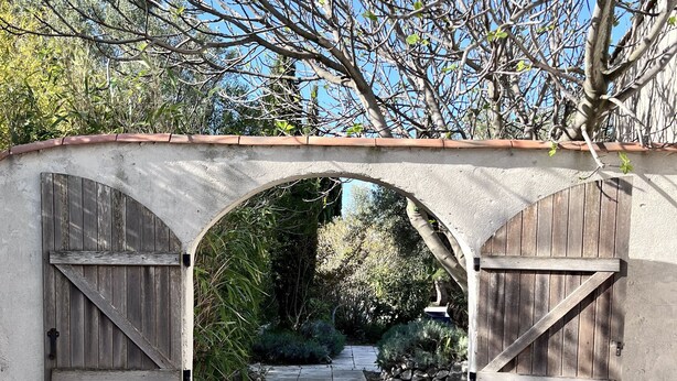 Un arc en pierre rustique, surmonté de portails en bois, mène à un chemin de gravier bordé de verdure.