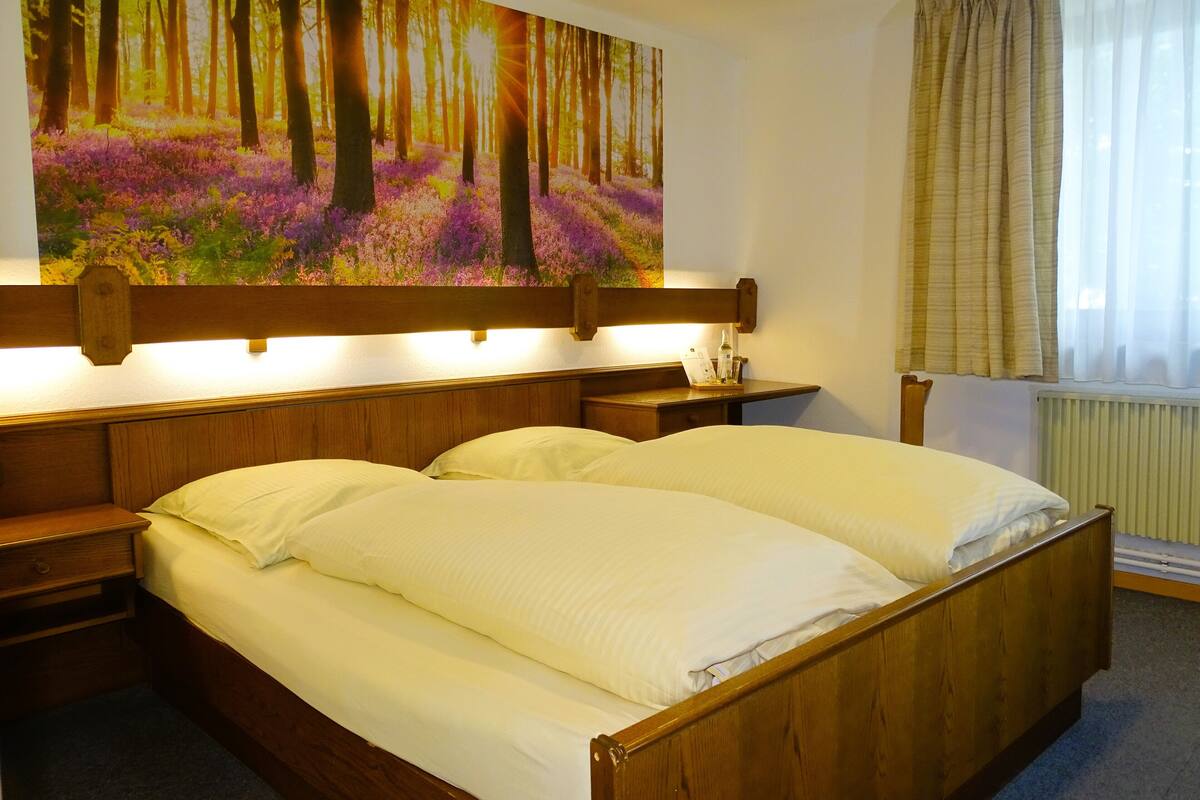 A double bed with wooden headboard, a nightstand with a lamp, and a painting of a forest scene above the bed.