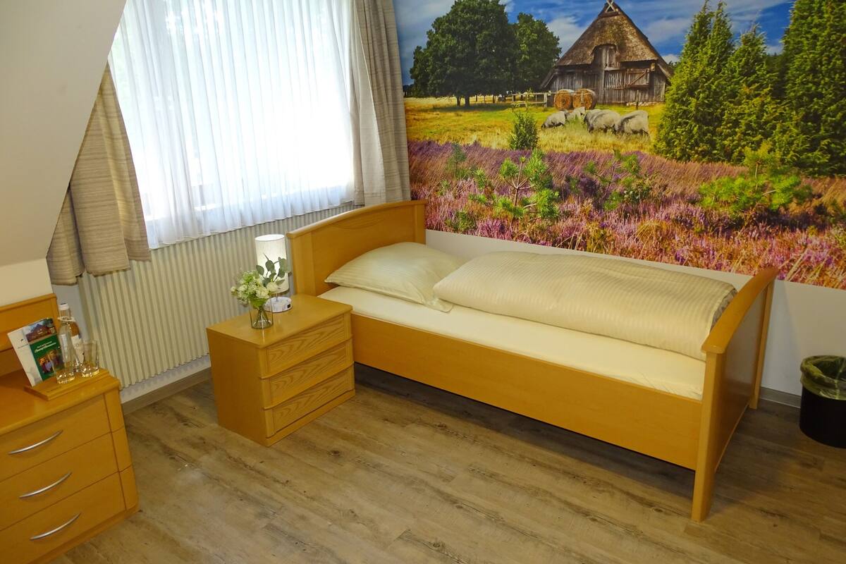 A bedroom with a wooden bed, a nightstand with a lamp, a dresser, and a window with curtains.