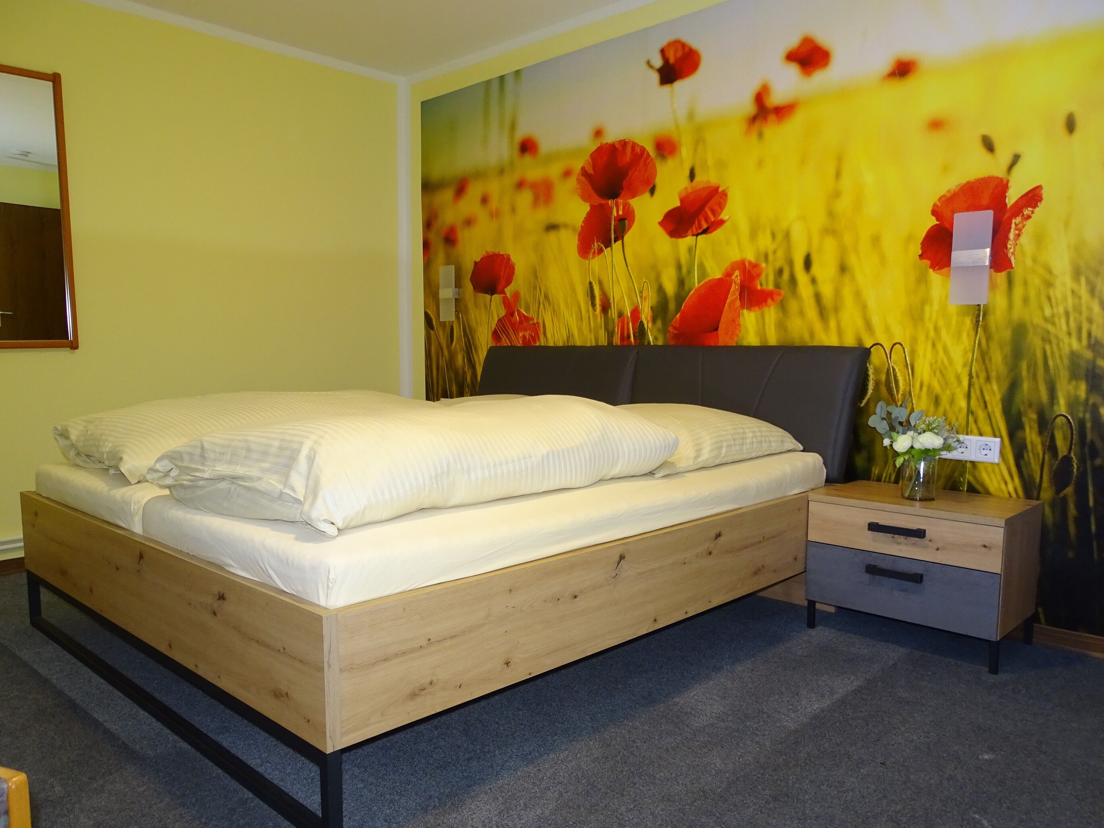 A bedroom with a wooden bed, a nightstand, a small drawer, and a wall mural of red flowers.