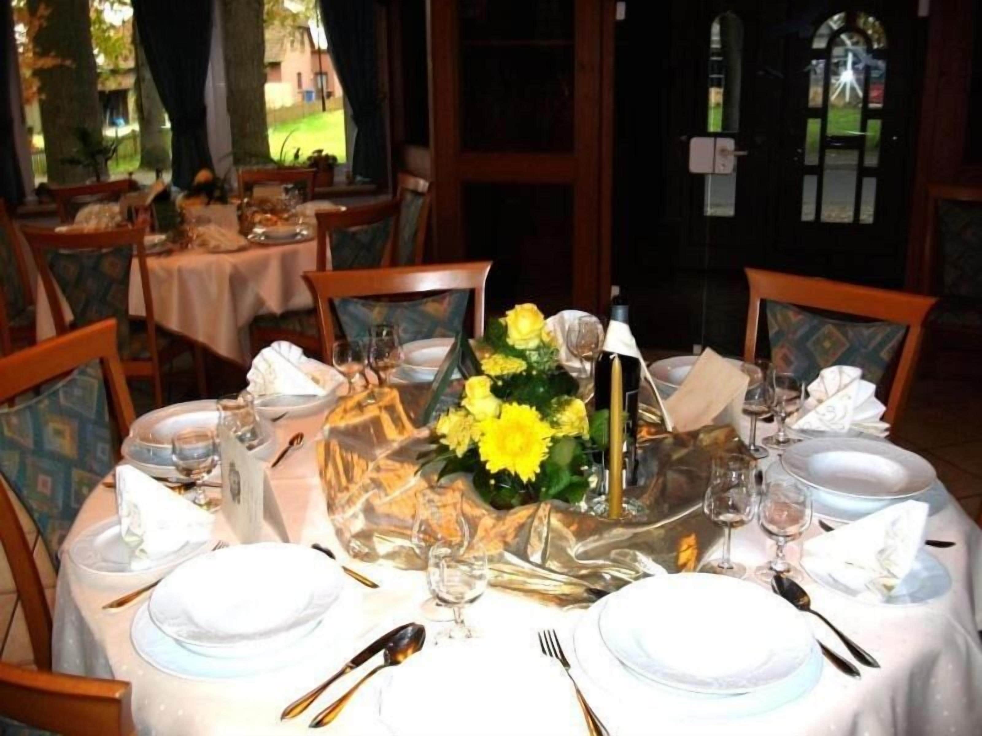 Una mesa de comedor preparada para una comida, con manteles blancos, platos, cubiertos y un centro de mesa con flores amarillas.