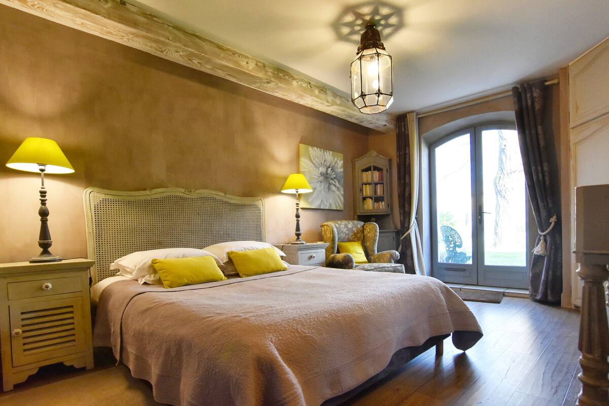 Un dormitorio con una cama grande, dos mesitas de noche con lámparas, una silla, una estantería y un ventanal con cortinas.