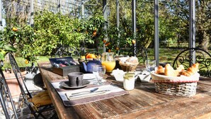 Un coin petit-déjeuner aménagé dans une véranda, avec une table, des aliments pour le petit-déjeuner et une vue sur les arbres et l’herbe.