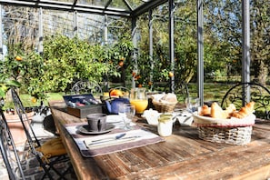 Desayuno preparado en un invernadero, con una mesa, alimentos para el desayuno y vistas a árboles y césped.