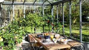 Un invernadero con una mesa de madera dispuesta para el desayuno, rodeado de plantas en macetas y vistas al jardín.
