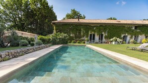 Una piscina rectangular con borde de piedra, rodeada por un césped cuidado y un edificio con techo de tejas y paredes cubiertas de hiedra.