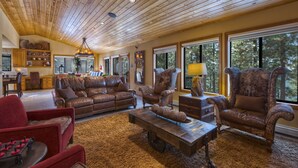 Ein geräumiges Wohnzimmer mit Holzbalkendecke, Ledersofas, einem Couchtisch und großen Fenstern mit Blick ins Freie.