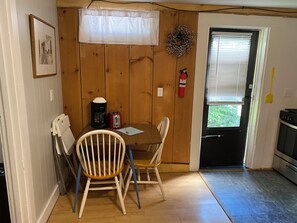 Una cocina pequeña con paredes de madera, una mesa de comedor con dos sillas, una cafetera, un tostador, un extintor y una ventana con persianas.