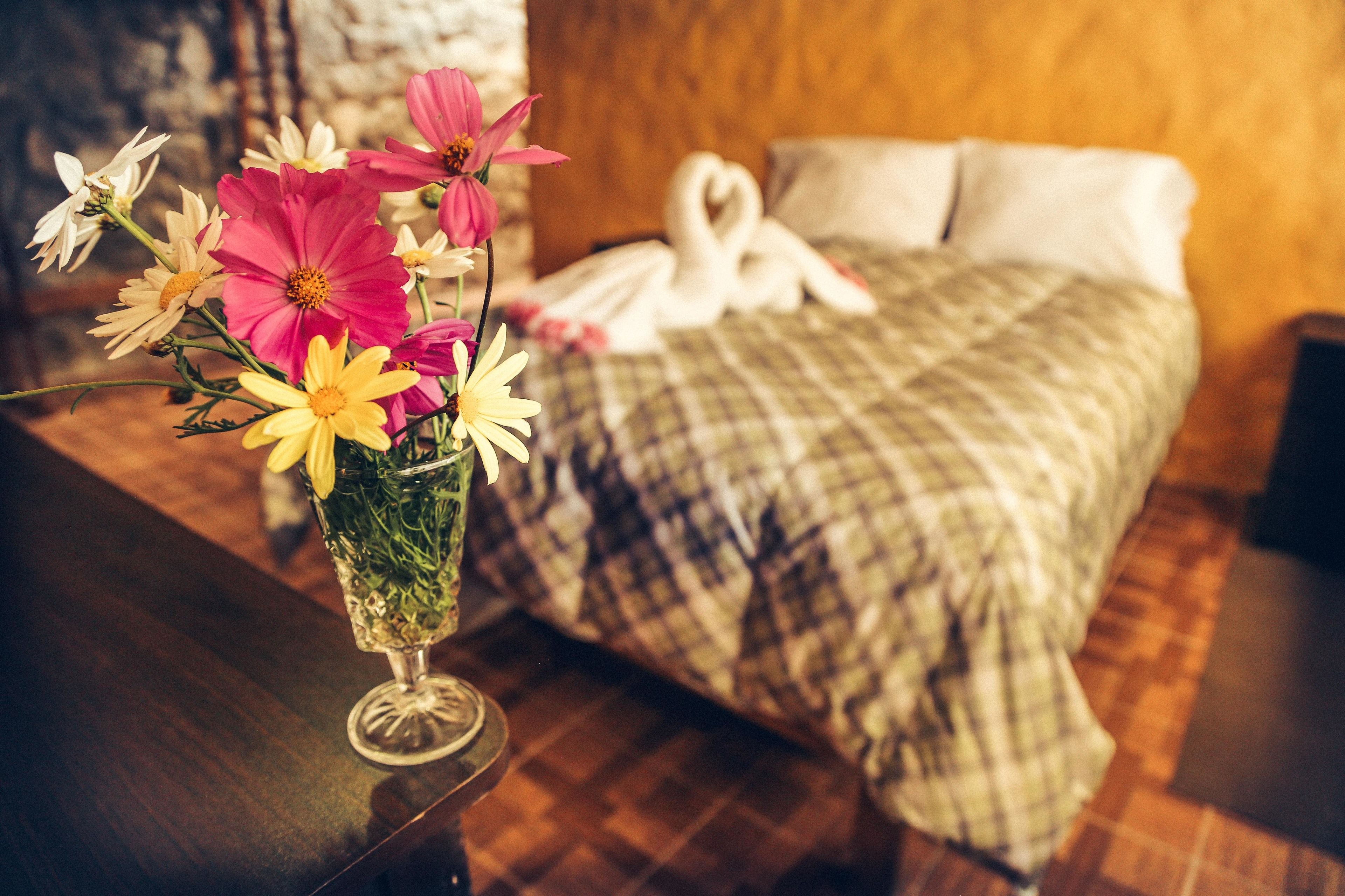 Una cama con un cojín en forma de corazón y un ramo de flores en un florero de vidrio sobre una mesa de madera.