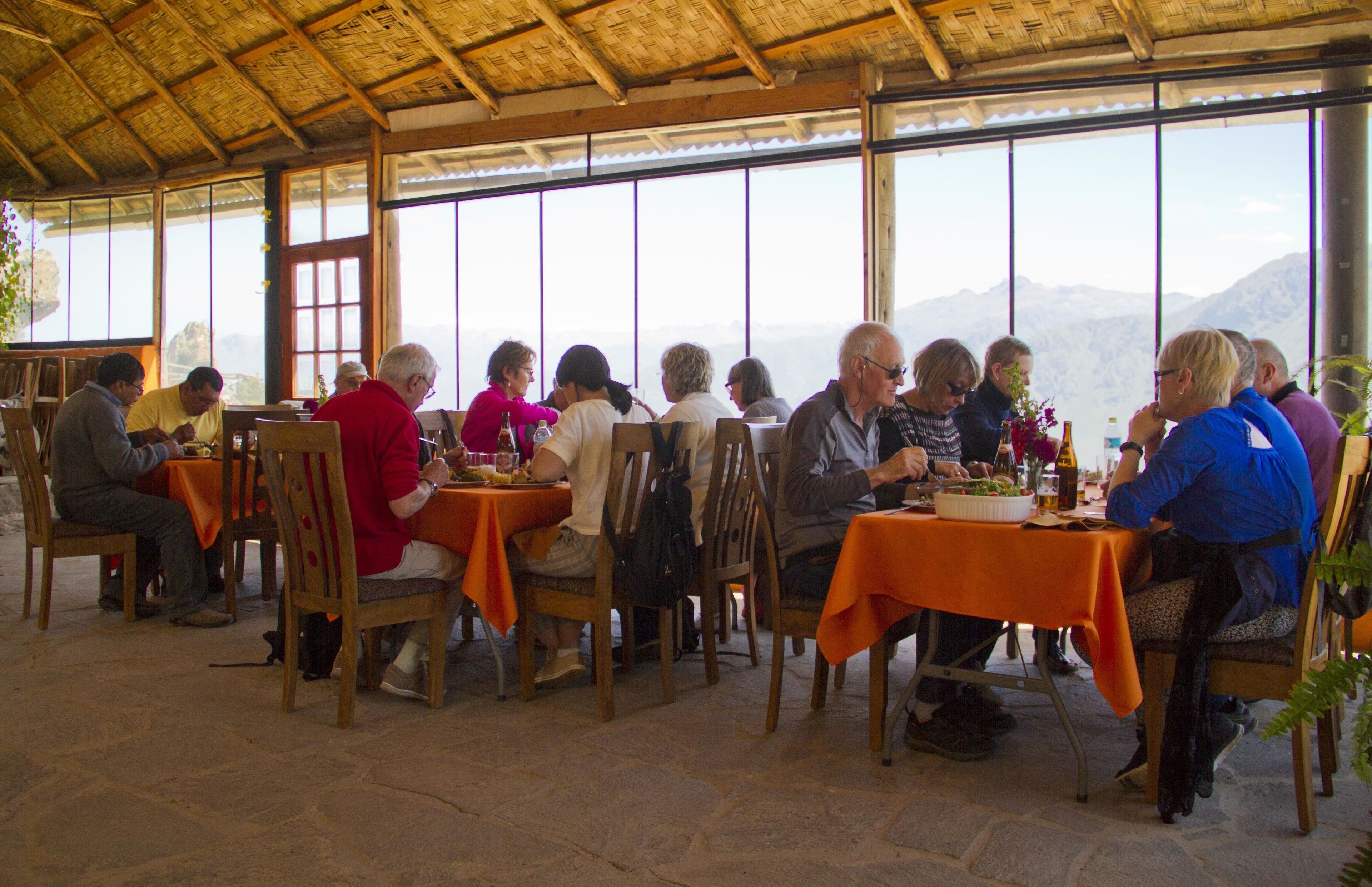 Een groep mensen dineert in een openluchtrestaurant met oranje tafellakens en houten stoelen, met een bergpanorama op de achtergrond.
