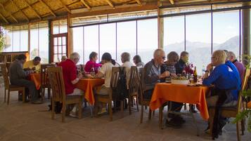 Een groep mensen dineert in een openluchtrestaurant met oranje tafellakens en houten stoelen, met een bergpanorama op de achtergrond.