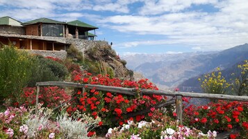 A mountain lodge with stone walls and a wooden structure, surrounded by vibrant red and pink flowers, and a view of distant mountains.