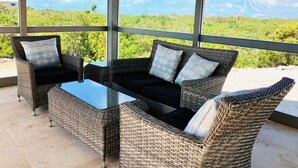 Un espacio al aire libre cubierto con mobiliario de mimbre y una mesa con cubierta de vidrio, con vistas a un paisaje exuberante.