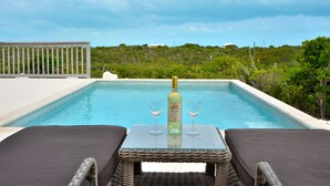 Escena junto a la piscina con una botella de vino y dos copas sobre una mesa, rodeada de sillones de descanso.