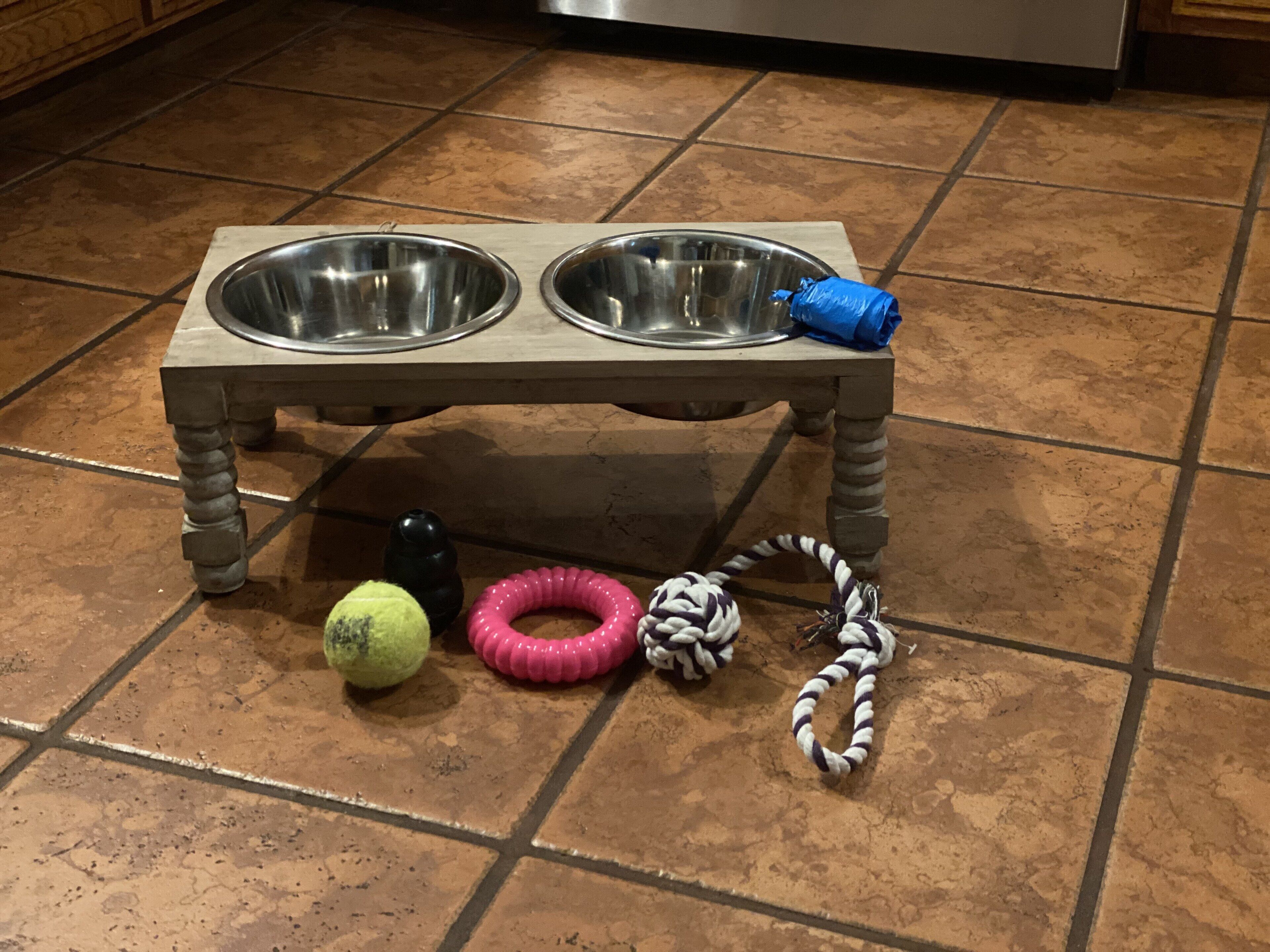 A pet feeding station with two bowls and a toy ball on a tiled floor.