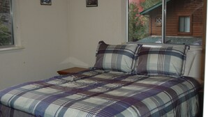 A bed with a plaid bedspread and two pillows, a window with a view of a cabin, and a small wooden nightstand.